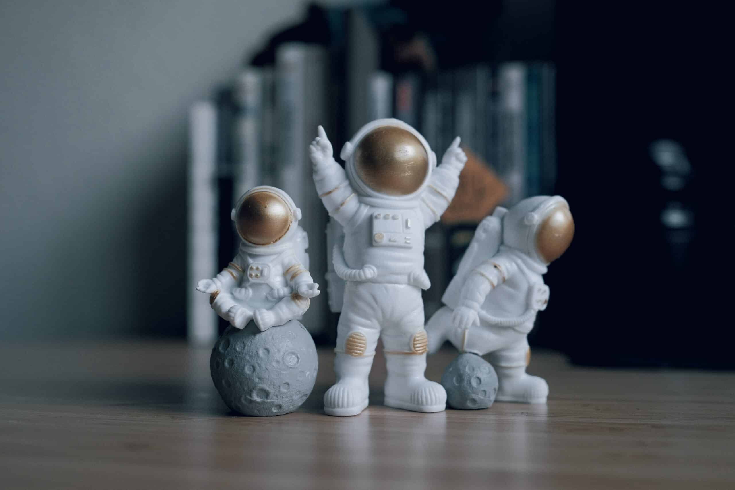 3 Astronaut figurines with one sitting on a moon, displayed on a desk with books behind them.